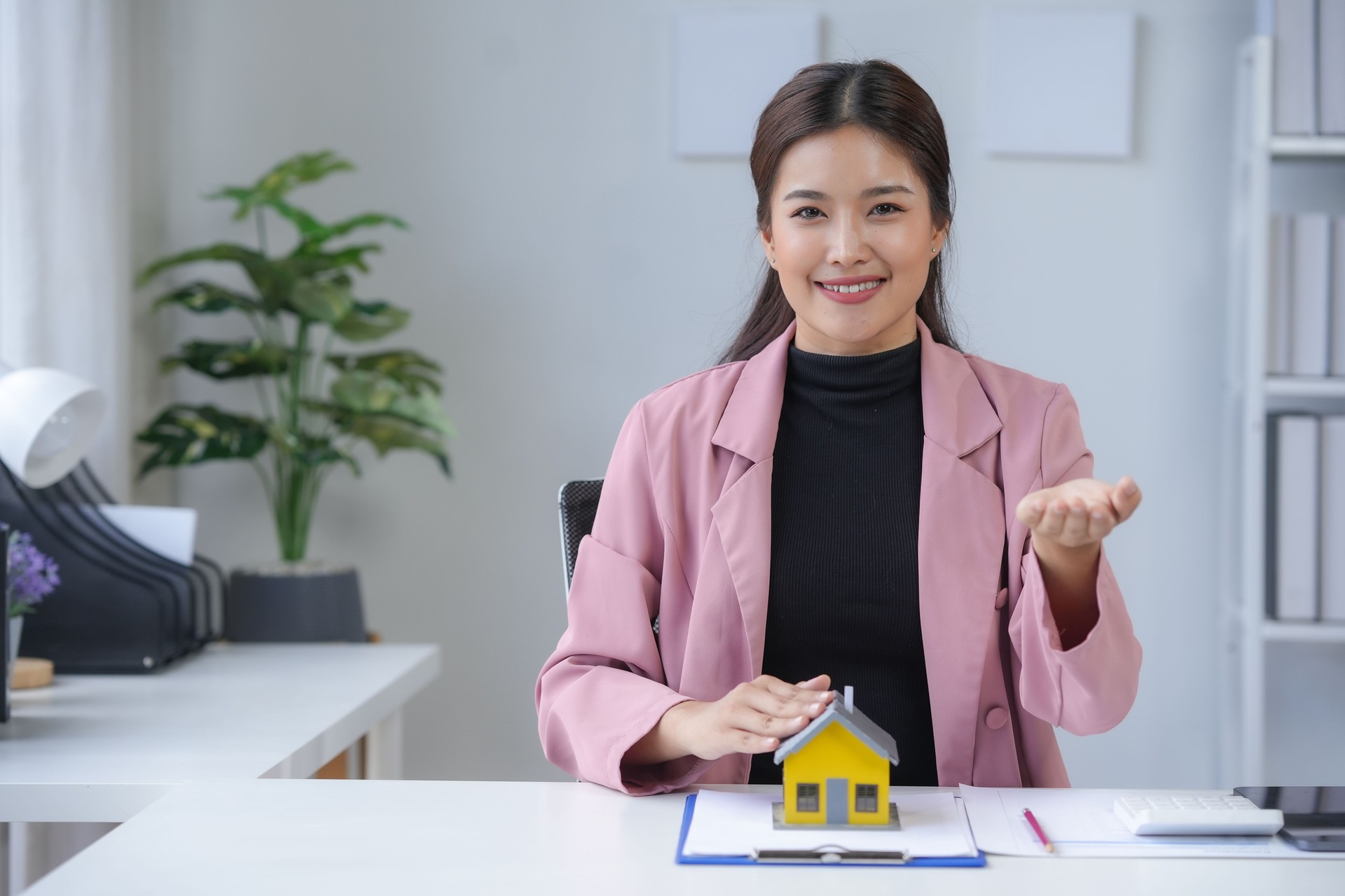 Mujer agente de bienes raíces presentando modelo de casa a la cámara y sonriendo en la oficina